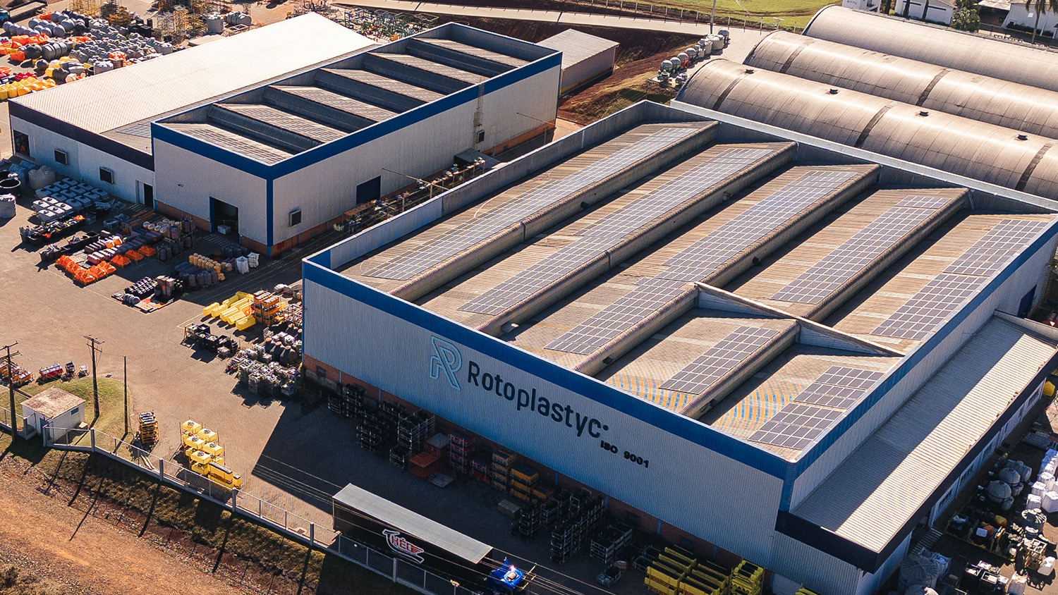 Vista aérea da unidade industrial da Rotoplastyc, com galpões, placas solares no telhado e peças rotomoldadas no pátio.